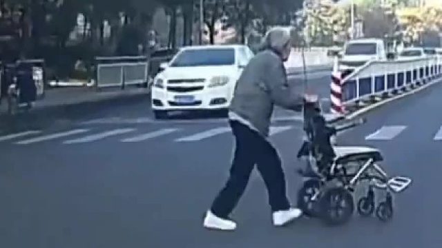 老奶奶推轮椅过马路被小车持续鸣笛吓到跌倒，艰难坐起来穿鞋