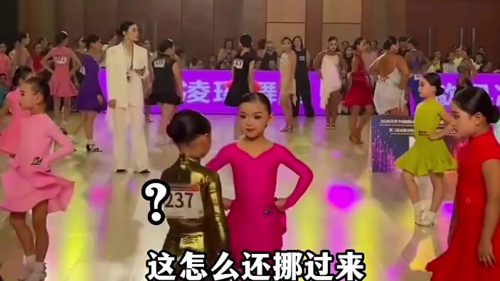 拉丁舞比赛现场粉衣小女孩站好后，另一个小朋友突然站女孩前面，两小朋友就开始面对面互相切磋舞姿