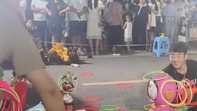 路人想用小圈去套大头玩偶，老板蹲在地上开心地笑了